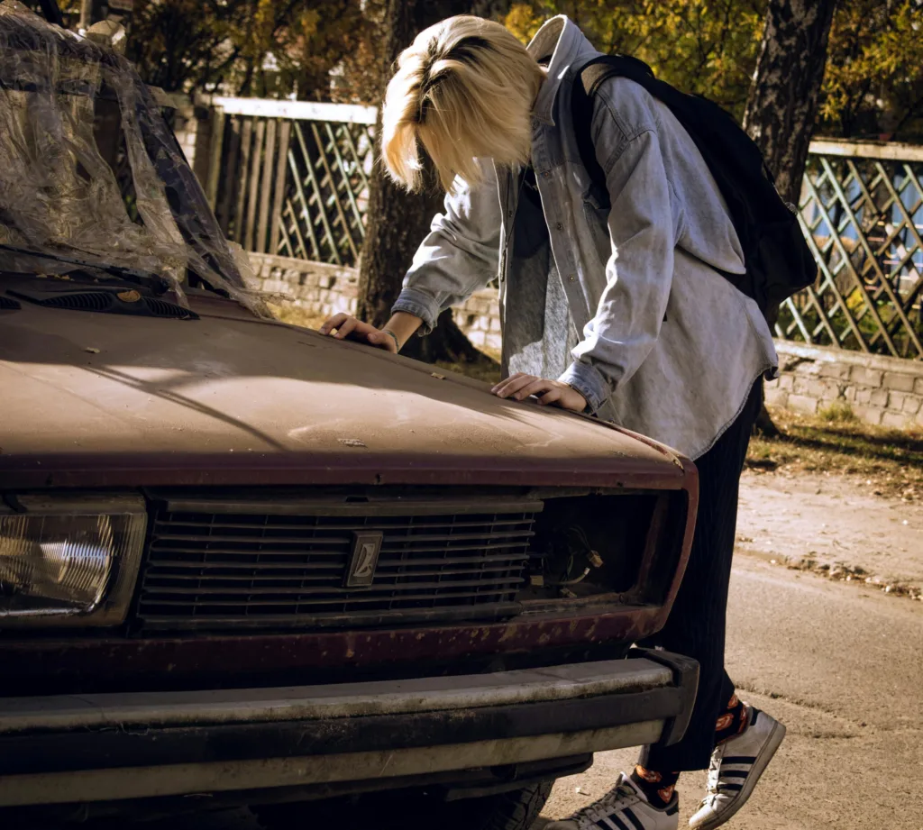 A young person leans on an abandoned car, reflecting stress and despair. Outdoors in natural daylight.
